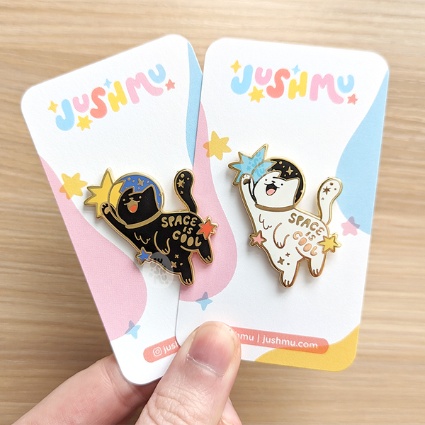 one black pin and one white cat pin that both say space is cool on them. Made by jushmu